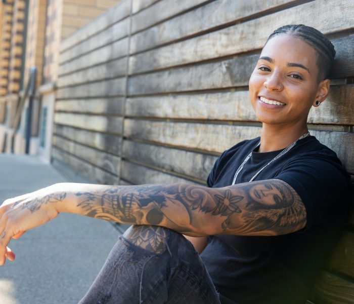 Young black woman with tattoos sits against wood wall.